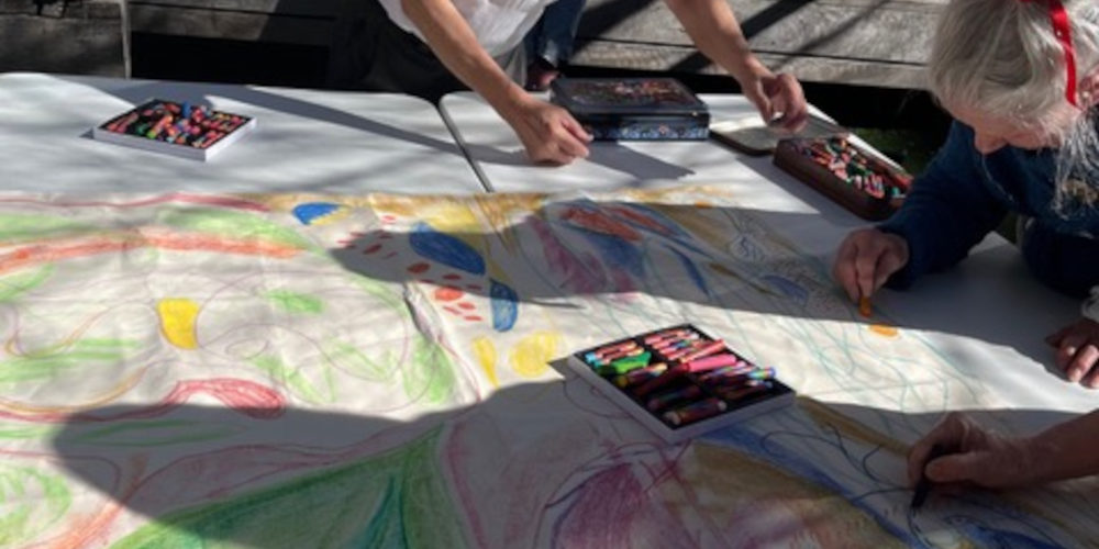 Many hands drawing with crayons and pastels on large pieces of white paper spread across many tables